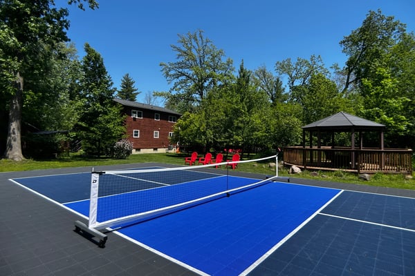 Blue tennis court with net in foreground, red Adirondack chairs, gazebo, and red wooden house surrounded by trees